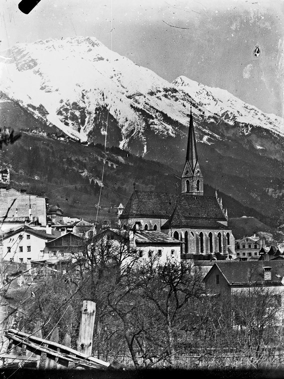 Blick auf die Pfarrkirche von St. Nikolaus und einige Wohnhäuser. Im Hintergrund kann man das Viadukt der Hungerburgbahn und die schneebedeckte Nordkette erkennen.
