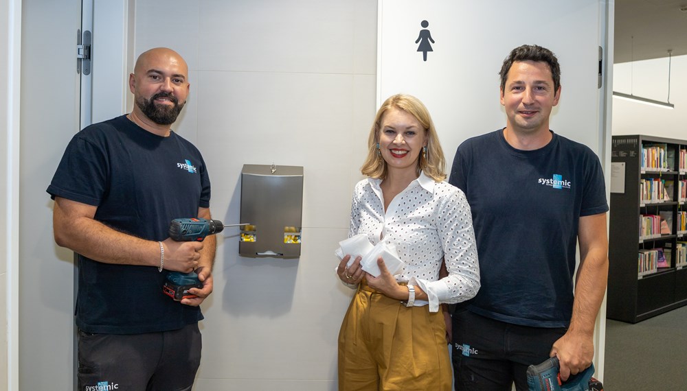 Vizebürgermeisterin Elisabeth Mayr mit den Mitarbeitern der Firma Systemic Reinigungstechnik, Ernad Cavkic und Hannes Müller (r.), bei der Installation eines Spenders für Menstruationsprodukte in der Stadtbibliothek.